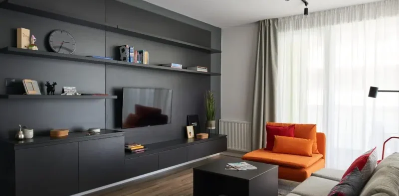 Világos, könnyed esésű függöny szűri a természetes fényt egy modern, belvárosi nappaliban a Corvin-negyedben. A letisztult vonalvezetés jól illeszkedik a sötét szekrényfalhoz és a színes ülőbútorokhoz.